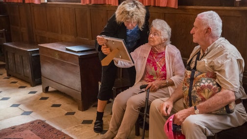 Two elderly visitors sitting on a bench in the Great Hall at Greyfriars being shown a photograph by a guide